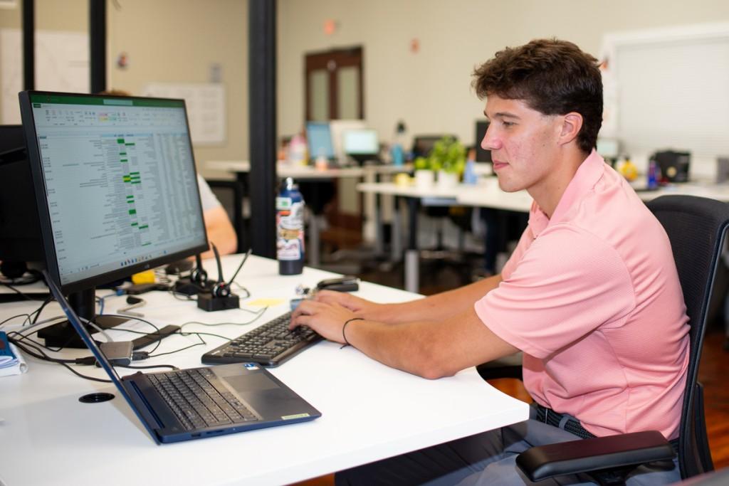 A student works at a desk with a computer displaying a spreadsheet in an office setting